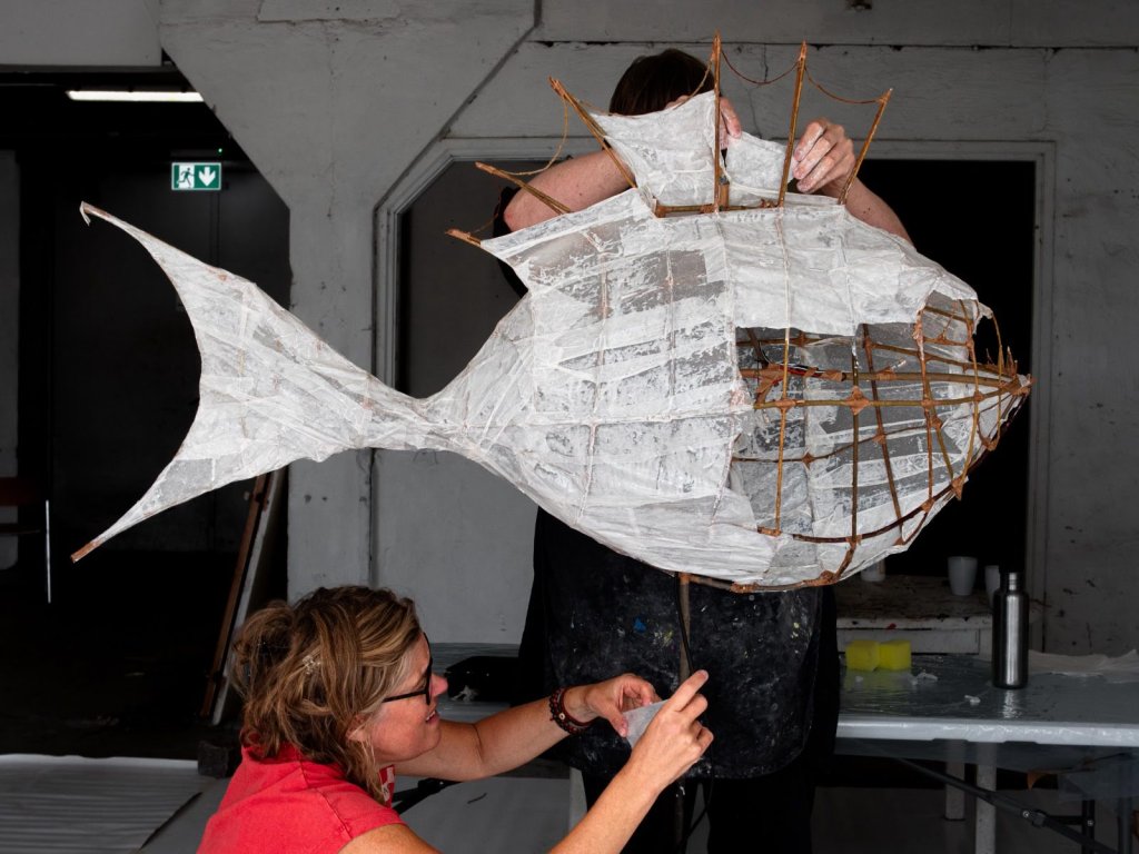 Två personer hjälps åt att bygga en stor fiskformad skulptur av papper och pilslanor.